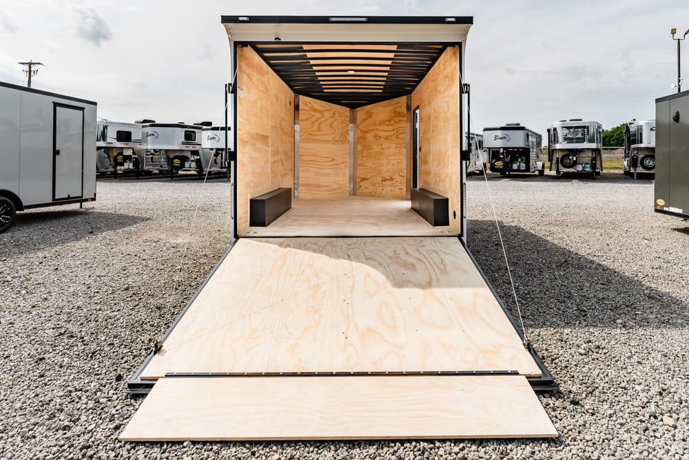 Empty open cargo trailer with ramp down, plywood interior, and two small benches on either side in a gravel lot with other trailers in the background in a dealership yard for loading.