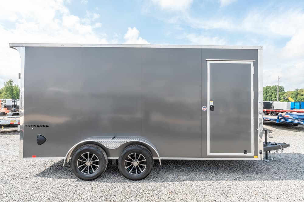 Gray enclosed cargo trailer with dual axles and diamond-plate fenders on a gravel lot, side view.