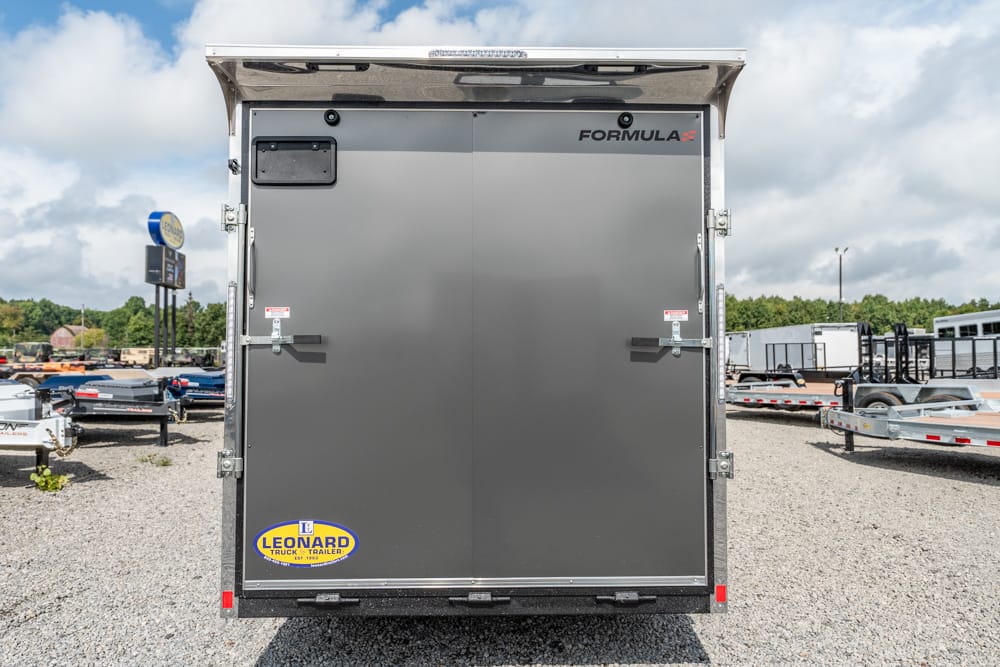 Rear view of a dark gray Formula enclosed trailer with two doors and metal latches, in a gravel trailer yard with other trailers nearby.