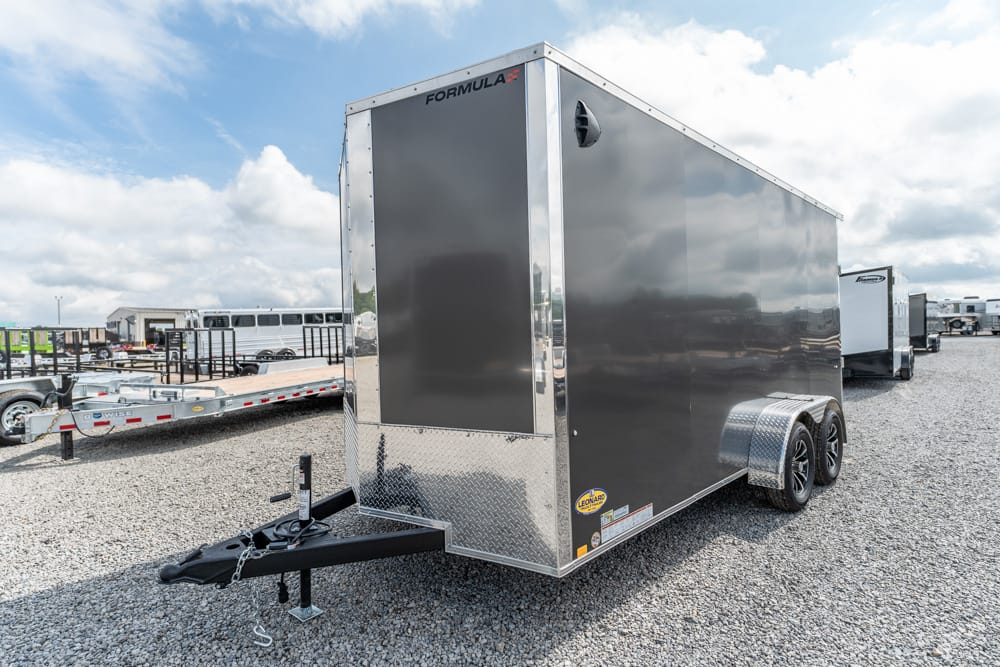 Grey enclosed cargo trailer with dual axles and diamond-plate front on a gravel lot, hitch at the front