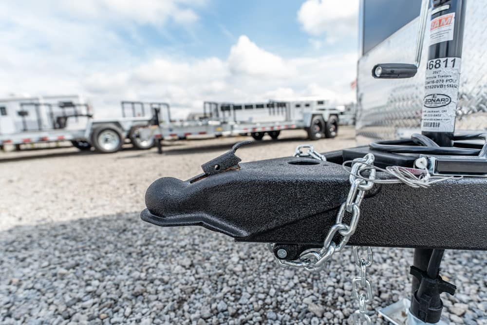 Close-up of a black trailer hitch with a steel safety chain on a gravel lot, trailers in the background.