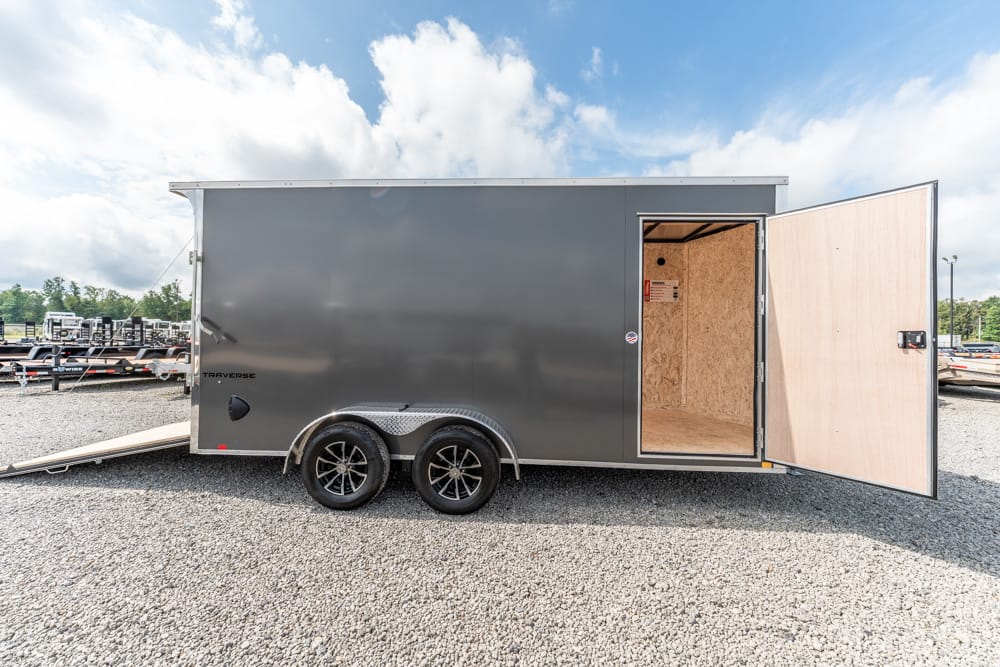 Gray two-axle enclosed cargo trailer on gravel, rear door open revealing plywood interior and a loading ramp nearby.