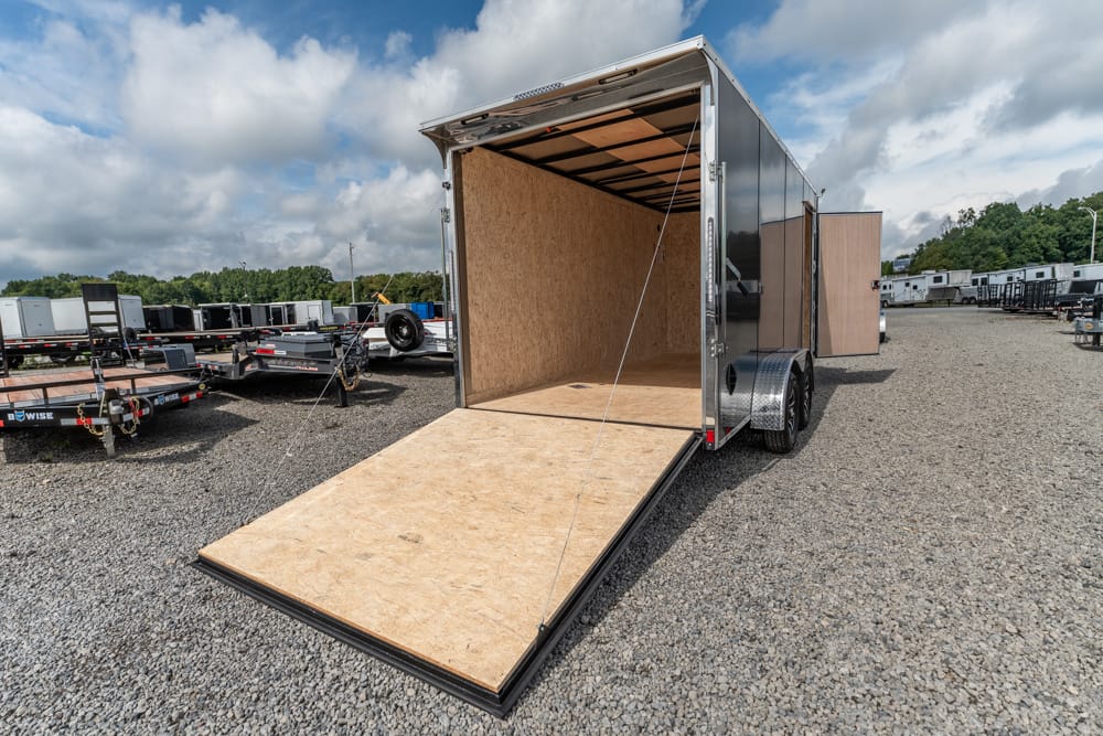 Open rear ramp of an empty enclosed cargo trailer in a gravel lot, exposing plywood interior and side door open behind.