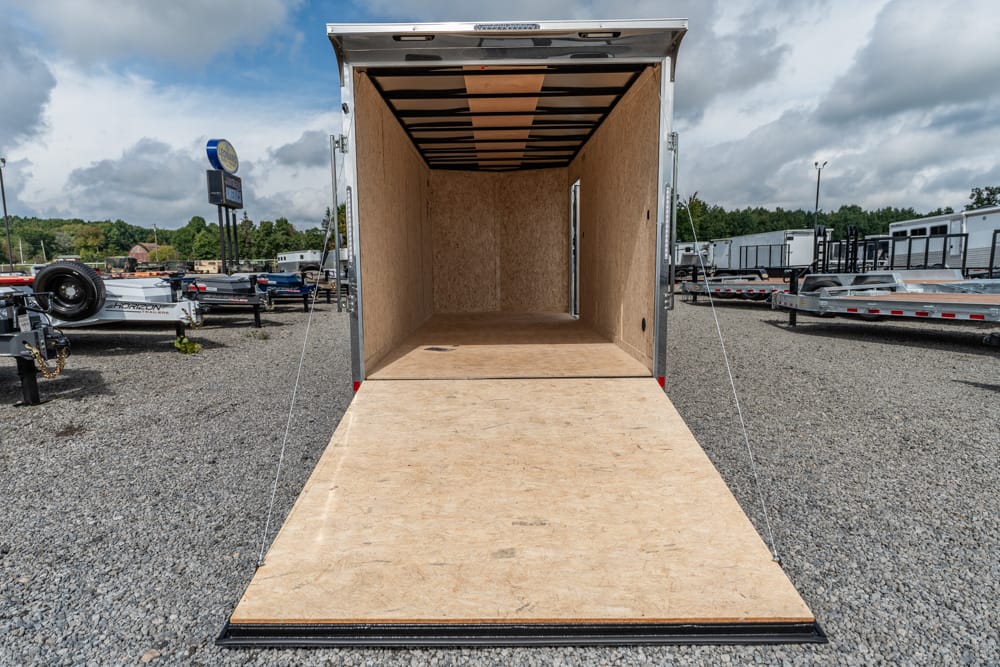 Open cargo trailer with the rear ramp lowered onto a gravel lot, showing plywood interior walls and ceiling.