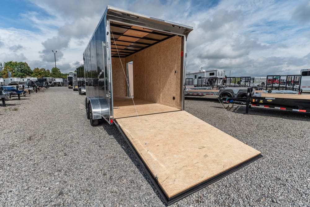 Open cargo trailer with the ramp down, showing plywood interior, on a gravel lot among other trailers.