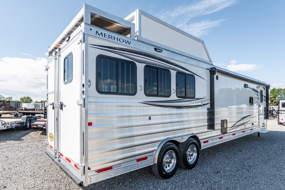 White Merhow horse trailer with tandem wheels parked on a gravel lot, side view, blue sky above.