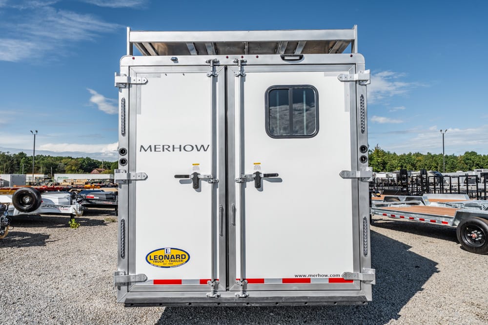 Rear view of a white Merhow horse trailer with a small window, Leonard trailer logo, in a gravel lot with other trailers nearby