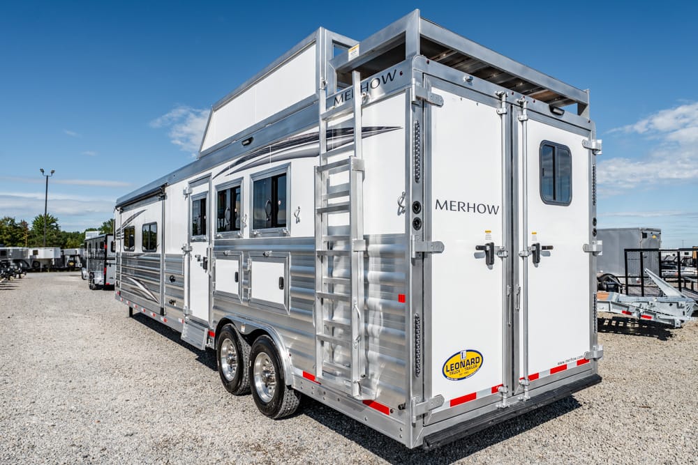 Silver Merhow horse trailer with dual rear doors, a side ladder, and windows on a gravel lot