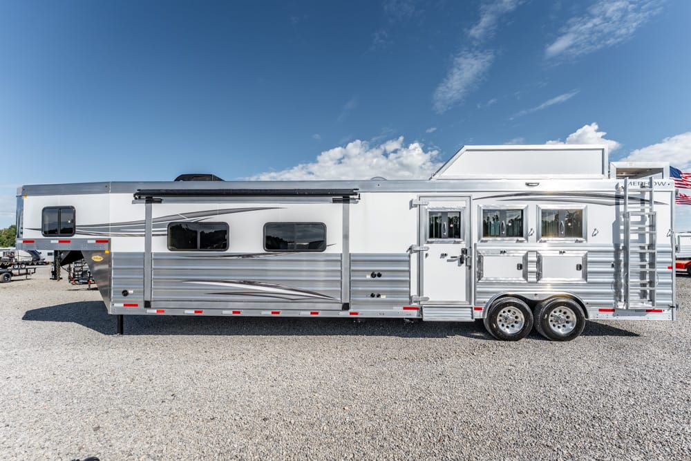 Long silver horse trailer with side windows, exterior storage, and a rear ramp on a gravel lot under a blue sky