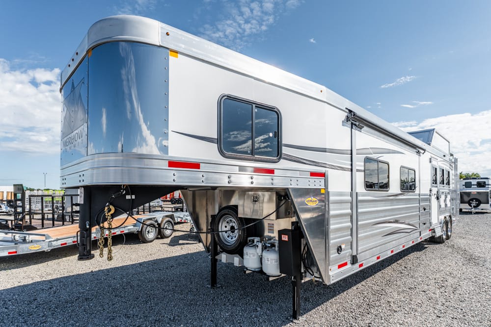 Gooseneck horse trailer with silver aluminum exterior, two axles, parked on a gravel lot with propane tanks at the hitch area.