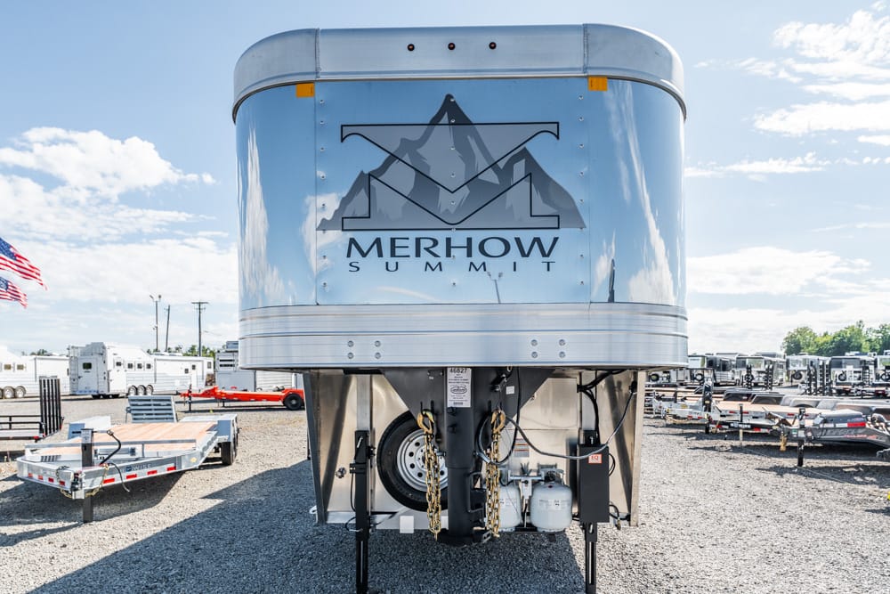 Front view of a silver Merhow Summit horse trailer with its logo on the front, parked in a gravel lot with other trailers nearby and an American flag in the background