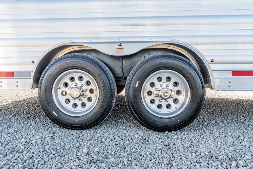 Twin axle trailer wheels with silver rims on a gravel surface.