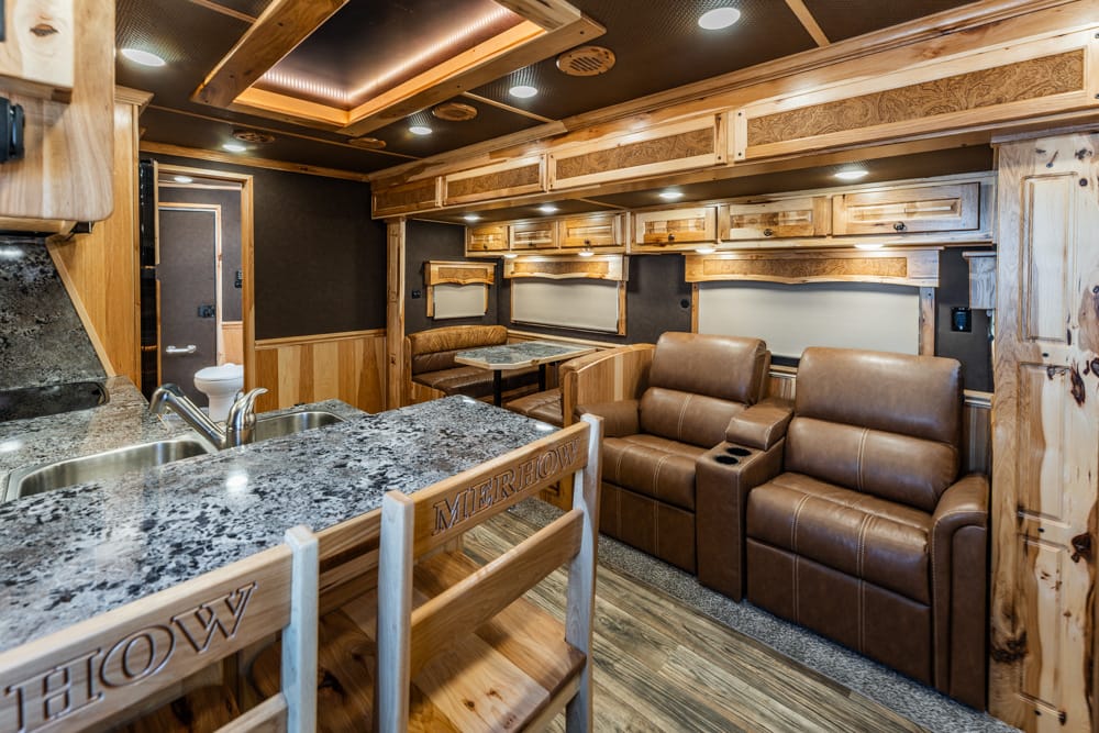 Interior of a wood-toned RV with granite kitchen counter, stools, and a leather seating area for relaxing and dining.