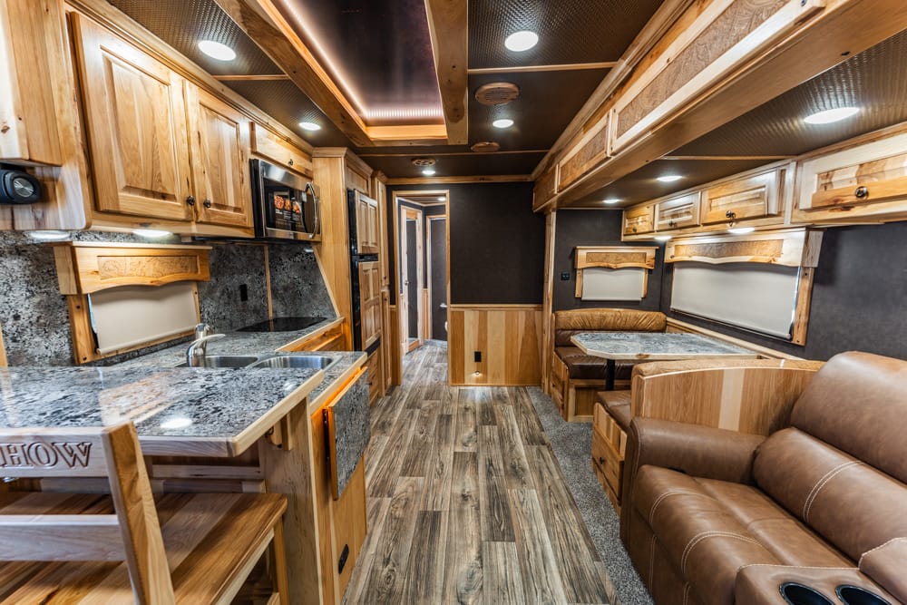 Interior of a wood-finished RV with a kitchen on the left, counter space, and a dining booth on the right; brown leather sofa nearby.