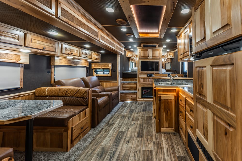 Warm, wood-walled RV interior featuring a leather sofa, dining booth, and a full kitchen with granite counters and upper cabinets