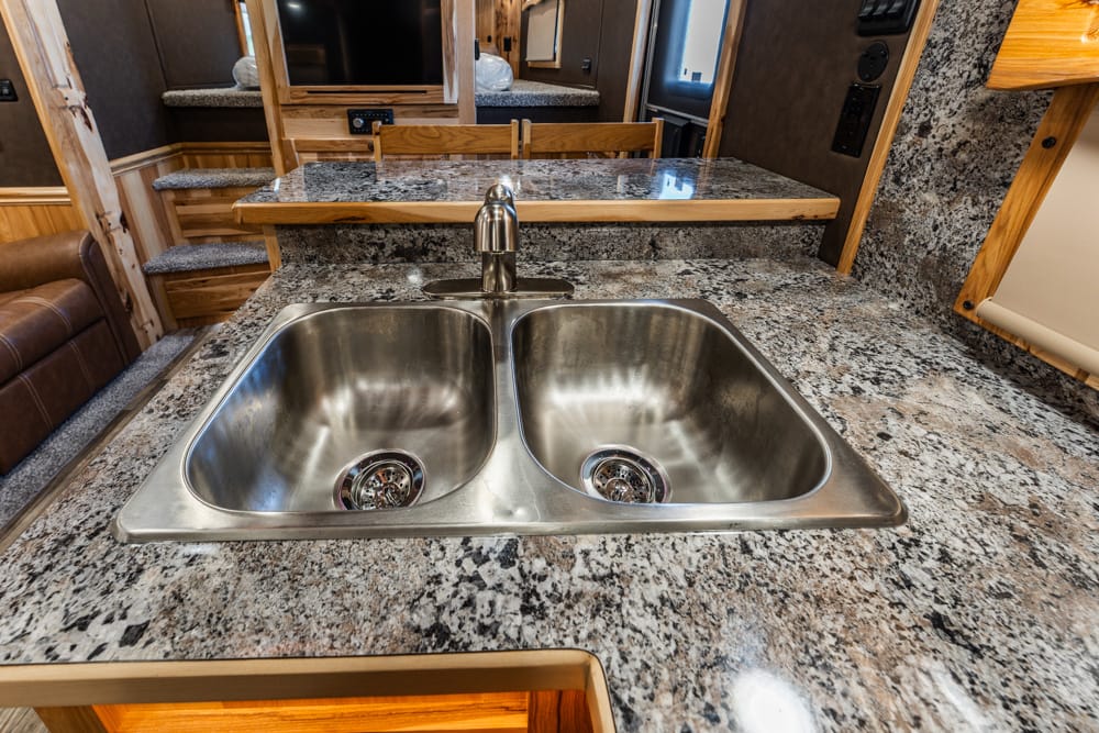 Double stainless steel, dual-basin kitchen sink with a chrome faucet on a speckled granite countertop inside an RV.”