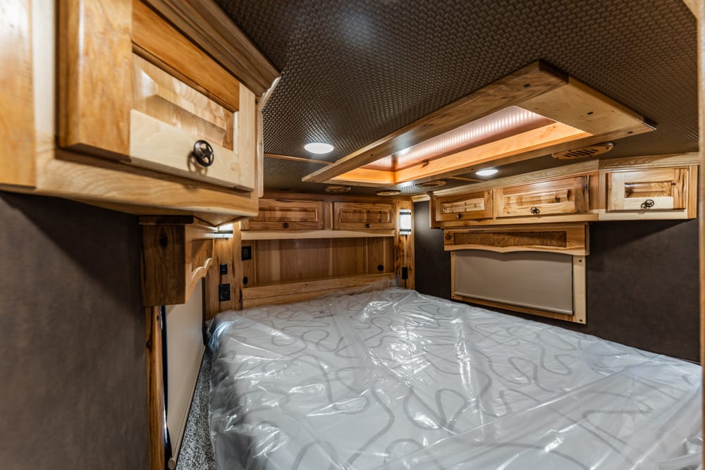 Wood-accented bunk area in a camper with overhead cabinets and a plastic-covered mattress.