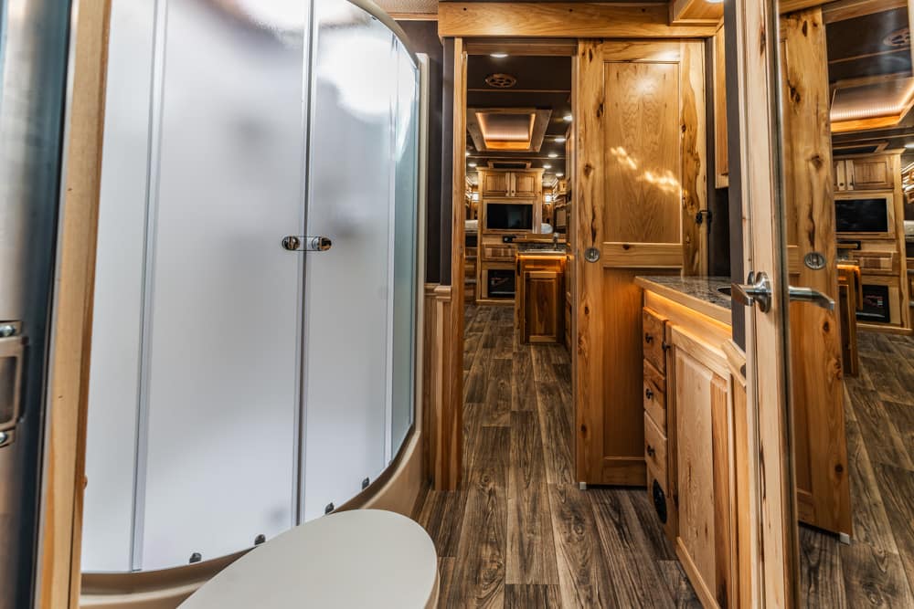 Wood-paneled RV bathroom with a curved frosted shower door, toilet nearby, and warm wood cabinetry along the hallway.
