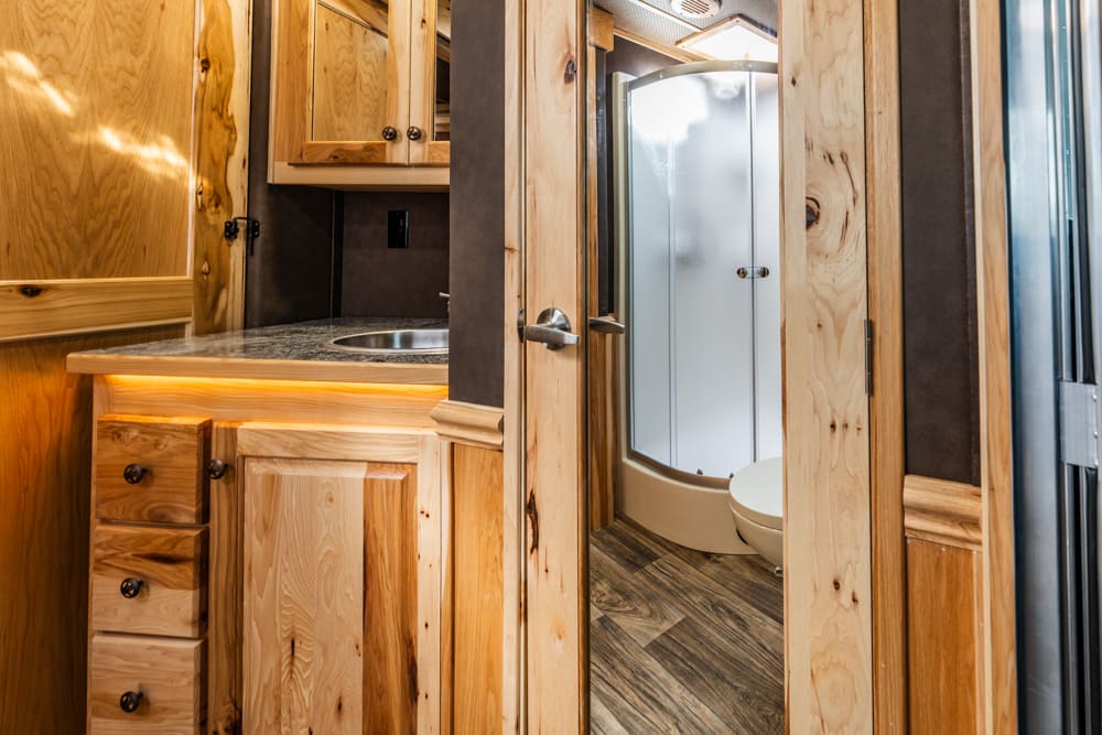 RV bathroom interior with warm wooden cabinets, granite countertop, and a curved shower enclosure visible through the doorway