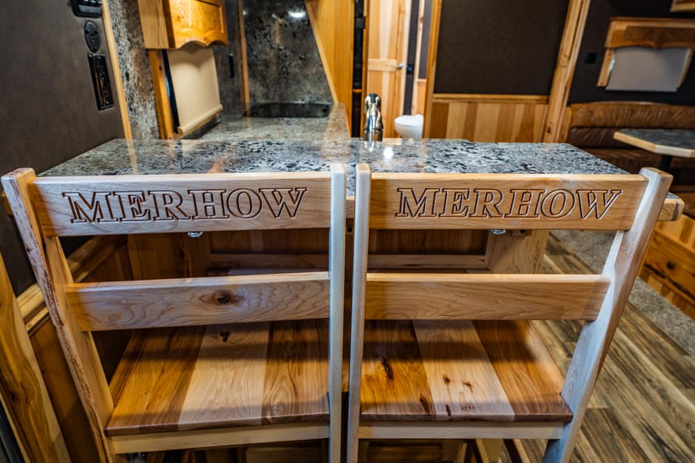 Two rustic wooden stools with engraved backrests reading MERHOW sit at a granite countertop in a wood-paneled kitchen. Prioritizes the chairs and the engraved branding in a warm, cabin-like setting.