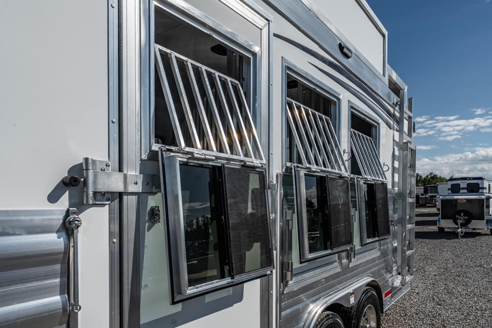 Silver horse trailer with open sliding windows and metal grilles along a gravel lot on a sunny day.