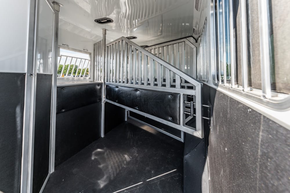 Interior of a metallic horse trailer showing a stall with a metal divider and black padded walls, with side windows along the length.