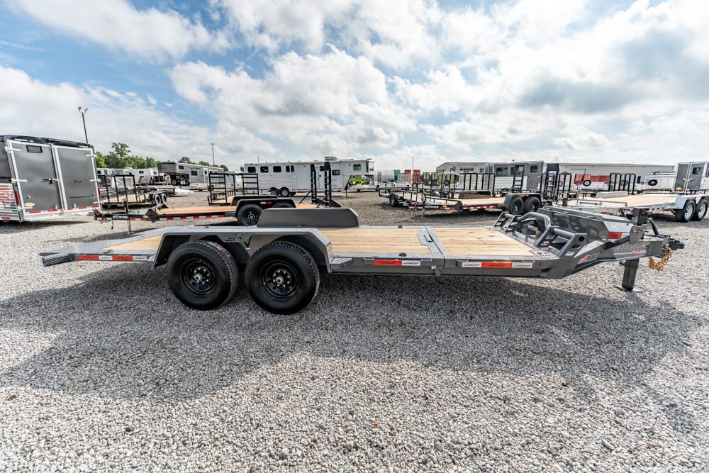 Flatbed trailer with dual axles and wooden deck parked on a gravel lot, with other trailers in the background. In foreground: a long utility trailer ready for hauling.
