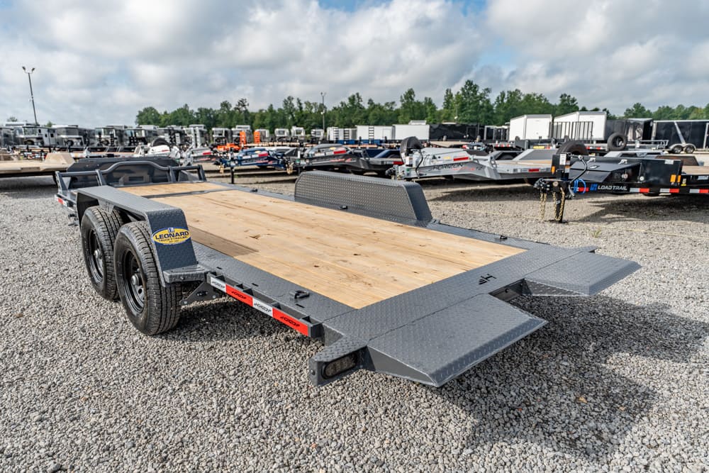Dual-axle flatbed trailer with a wood deck and fold-down ramps on a gravel lot, surrounded by other trailers.