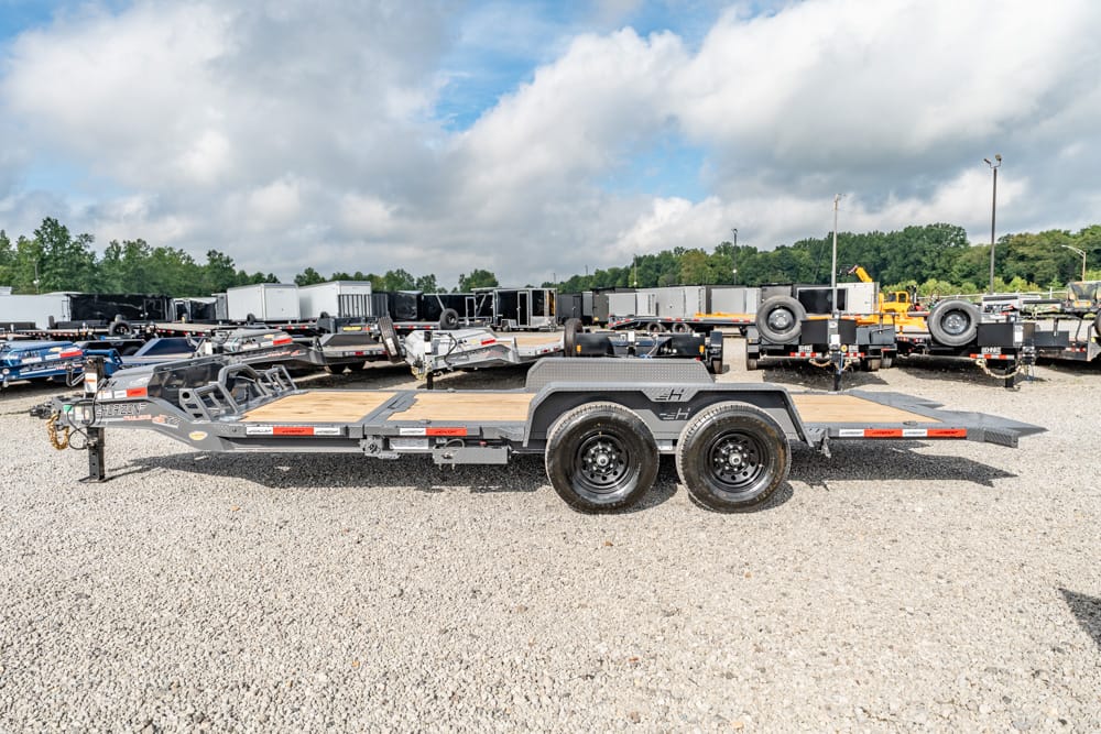 Dual-axle flatbed trailer with wooden deck parked on a gravel lot among other trailers, trucks, and equipment in the background, sunny with clouds.