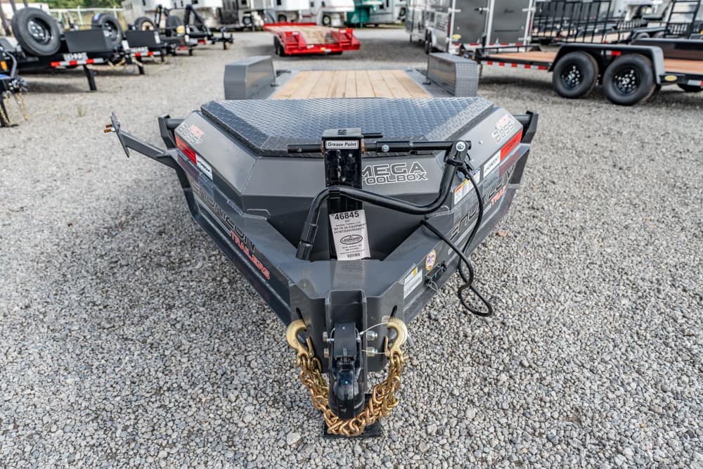Front view of a gray Mega Tool Box utility trailer on a gravel lot, showing the hitch, safety chains, and labels.