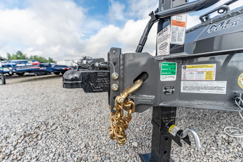 Close-up of a trailer hitch with a gold safety chain and padlock, attached to the coupler on a gravel lot with parked cars in the background.