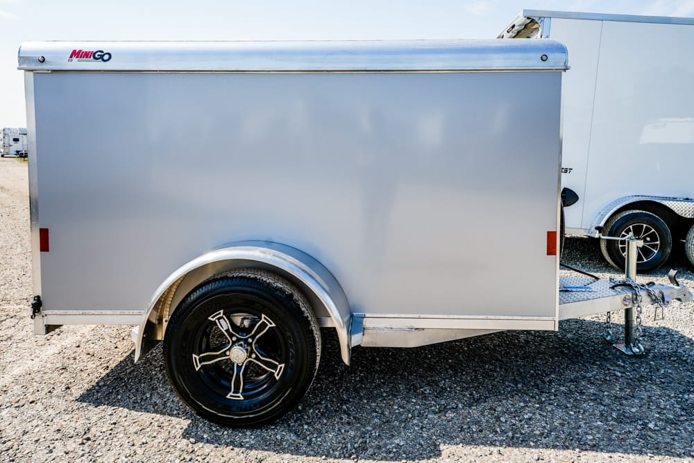 Silver enclosed trailer on a gravel lot, attached to a trailer hitch; single black rimmed wheel visible; MINIGO logo on upper front edge.