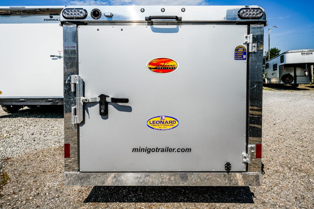 Rear of a silver trailer door with Sundowner and Leonard logos and minigot trailer.com on a gravel lot.