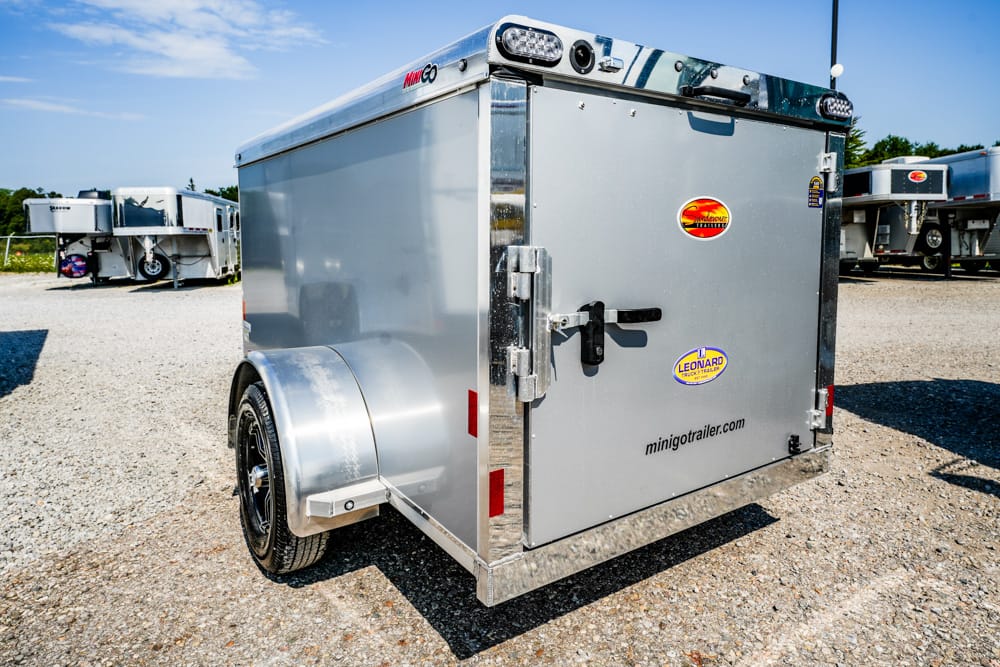 Silver enclosed single-axle trailer with a rear double-latch door and fender, parked on a gravel lot with other trailers nearby.