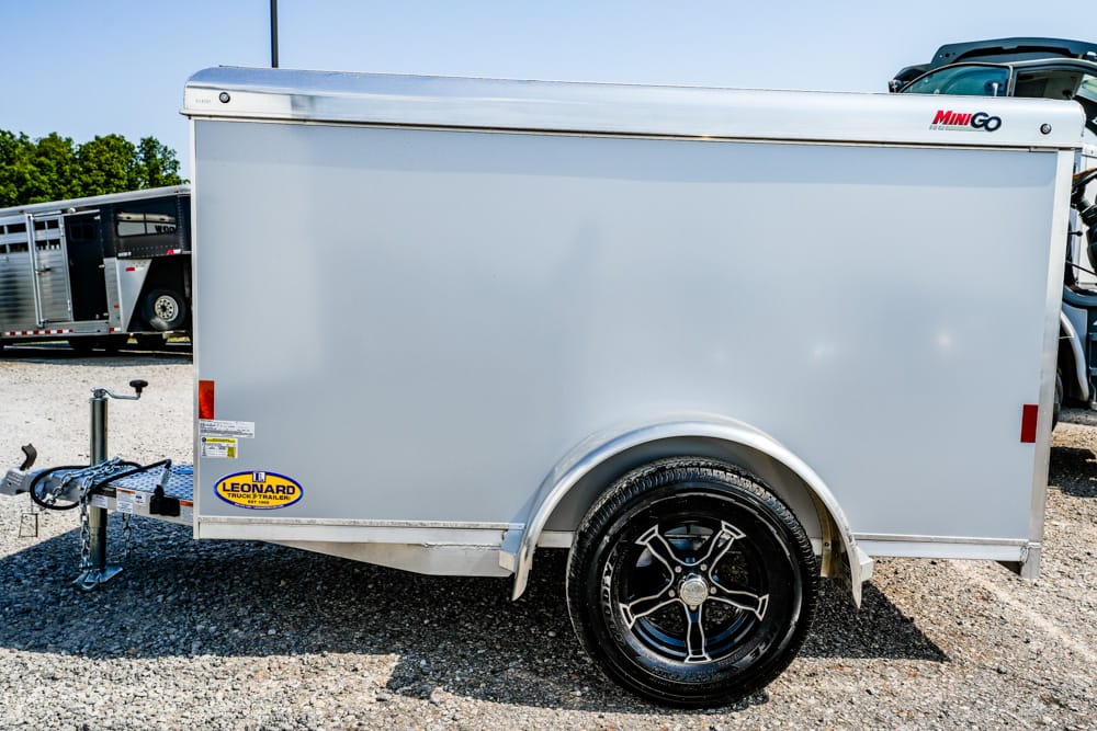 Silver enclosed cargo trailer with black alloy wheel and curved fender, hitched to a trailer tongue in a gravel lot.