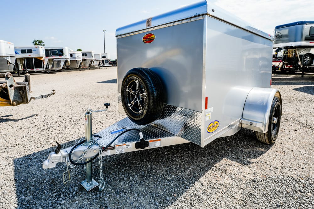 Silver enclosed utility trailer with a mounted spare tire, hitch, and jack in a gravel trailer yard.