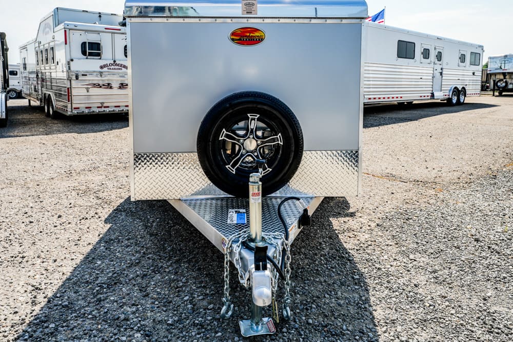 Front view of a silver trailer with a mounted spare tire and hitch, on a gravel lot with other trailers in the background, yard wide.