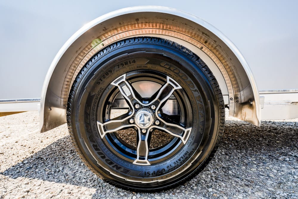 Close-up of a black and silver alloy wheel inside a curved metal fender on a trailer, resting on gravel.