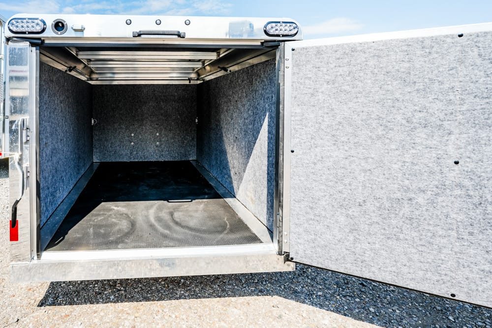 Open empty cargo trailer interior with gray, fabric-covered walls and a black non-slip floor, set on gravel under a blue sky.