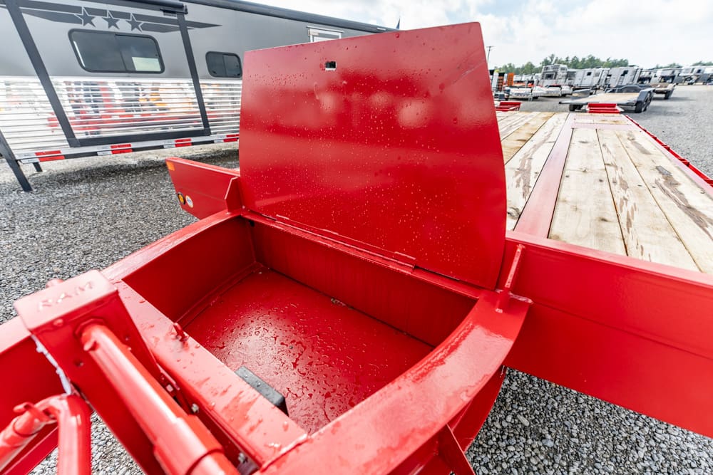 Close-up of a red metal hopper on a flatbed trailer, empty, with a wooden deck and gravel lot in the background.