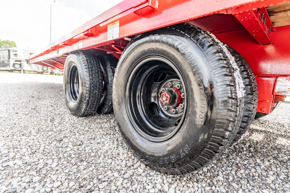 Red trailer with heavy-duty black tires parked on gravel; close-up of wheel and axle area with red lug nuts visible.