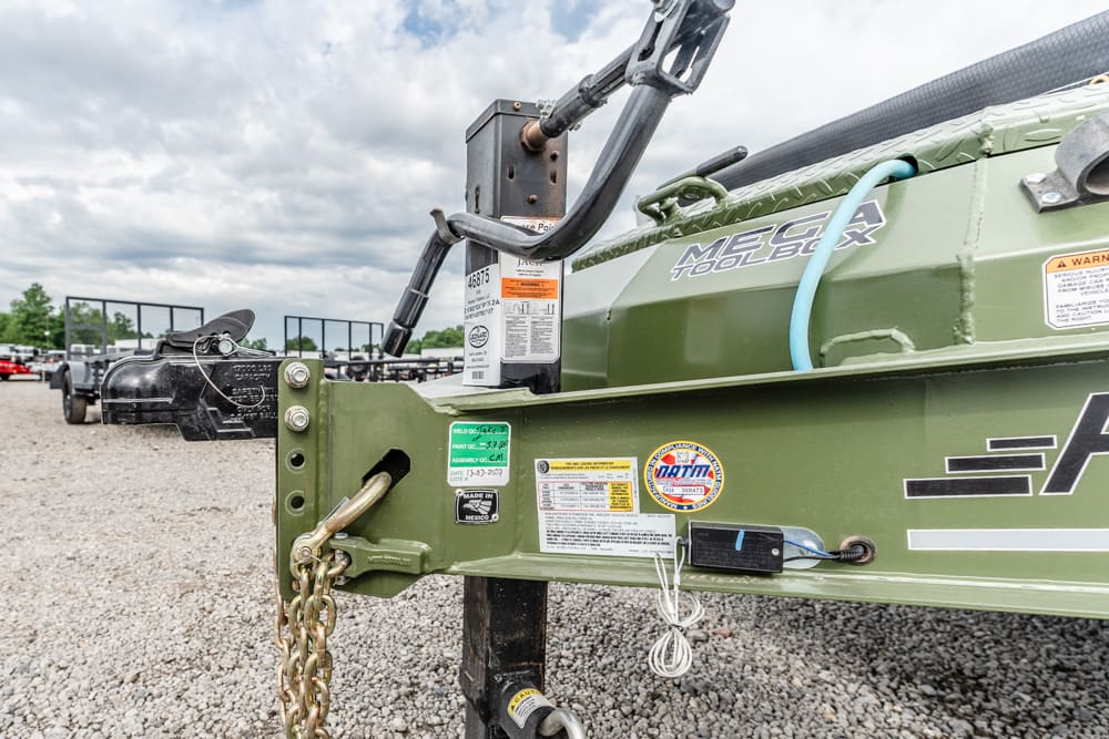 Close-up of a green utility trailer hitch with safety chains on a gravel lot; a black hitch mechanism is in the background with other trailers visible behind.