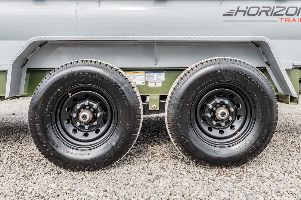 Dual-axle trailer wheels with black rims on a gravel surface, showing lug nuts and hub assemblies.