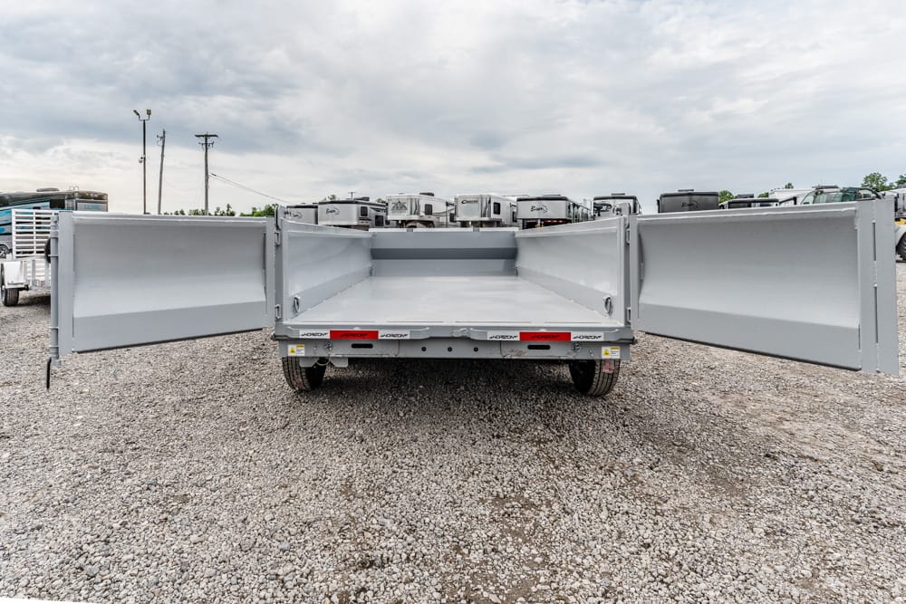 Open-back dump trailer with wide side doors swung open in a gravel lot, showing an empty cargo bed against a cloudy sky.