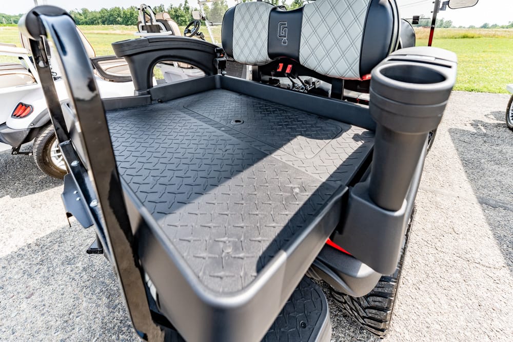 Close-up of the cargo area of a black golf cart with a diamond-plate bed, gray quilted seats, and cup holders, on a sunny course.