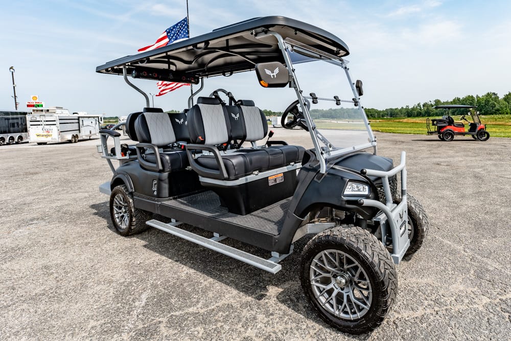Six-seat rugged golf cart with a sun canopy and large off-road tires parked on a gravel lot, American flag attached above.
