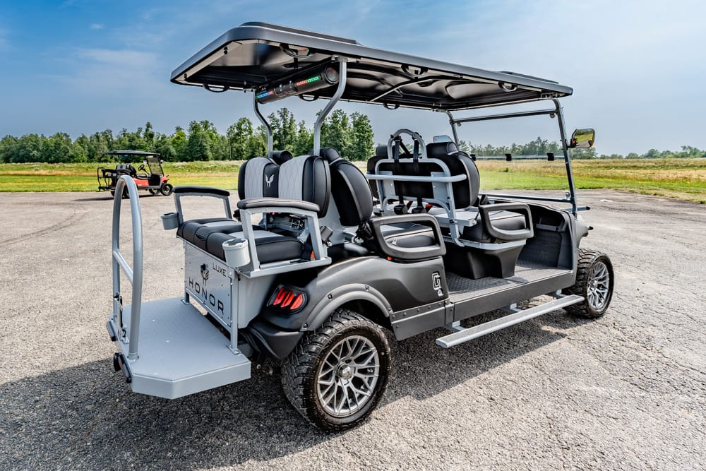 Gray four-seat golf cart with a canopy and rear access platform, parked on a paved lot with a field in the background.
