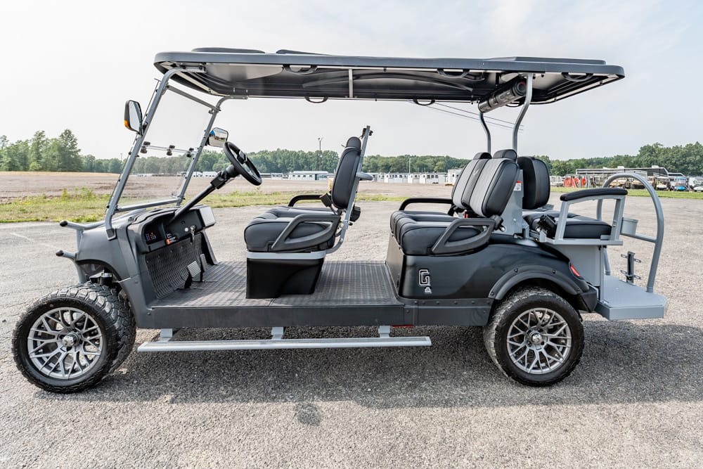 Six-passenger gray shuttle-style golf cart with a roof, open sides, and large off-road tires parked on gravel.