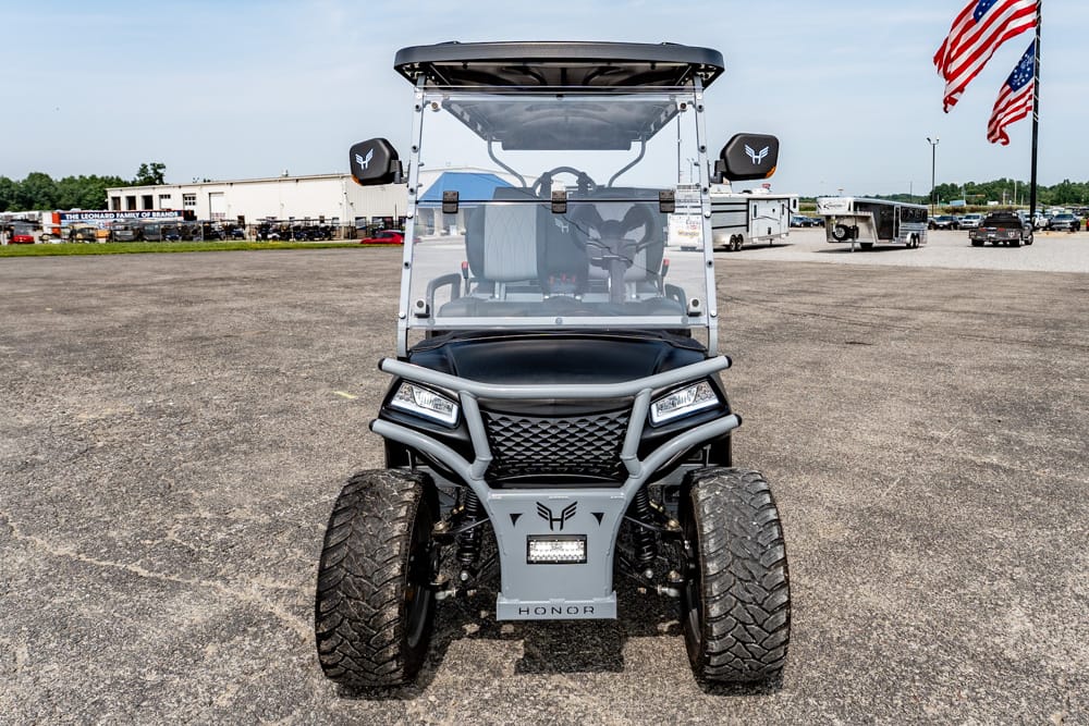 Front view of a gray off-road utility cart with large tires, windshield, and protective bumper, parked in a paved lot.
