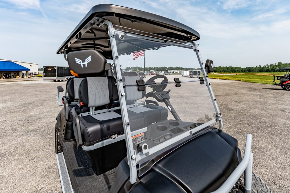 Front-right view of a four-seat golf cart with a clear windshield, grey seats, and a black roof, parked in a paved lot with an American flag in the background.